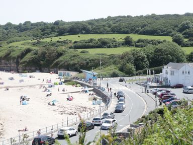 A beach with people and umbrellas next to a road with parked cars and green hills in the background at Ty Gilbert in Benllech