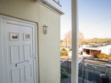 A white front door with a wall-mounted light next to a balcony with glass and metal railings overlooking a street and hills at 1 Lyndhurst in Salcombe