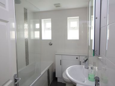 A bathroom with a bathtub shower, two small windows, a sink, and a cabinet at 1 Lyndhurst in Salcombe