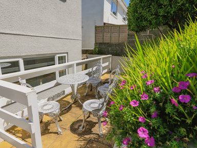 A small outdoor patio with white metal table and chairs next to a white building with windows and purple flowers at 1 Lyndhurst in Salcombe