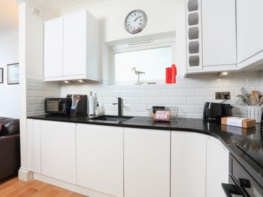 A kitchen with white cabinets black countertop a microwave toaster and a clock above the window at 1 Lyndhurst in Salcombe