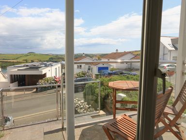 A balcony with wooden table and chairs overlooking a street with houses and green hills at 1 Lyndhurst in Salcombe