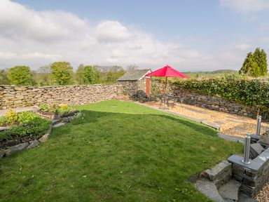 A garden with a grass lawn stone walls a small shed a red patio umbrella with a table and chairs at Rose Cottage in Lowick Bridge
