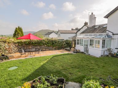 A garden with a green lawn a patio table with a red umbrella and chairs next to a white house with a glass conservatory at Rose Cottage in Lowick Bridge