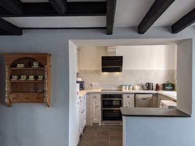 A kitchen with white cabinets black oven and hob wall-mounted wooden shelf with cups and bowls at Rose Cottage in Lowick Bridge