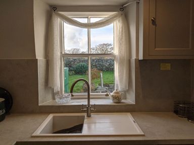 A kitchen sink under a window with sheer curtains and a teapot on the windowsill at Rose Cottage in Lowick Bridge