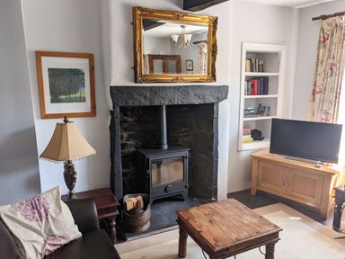 A living room with a wood-burning stove in a stone fireplace a wooden coffee table a TV on a wooden cabinet a lamp on a side table and a sofa with cushions at Rose Cottage in Lowick Bridge