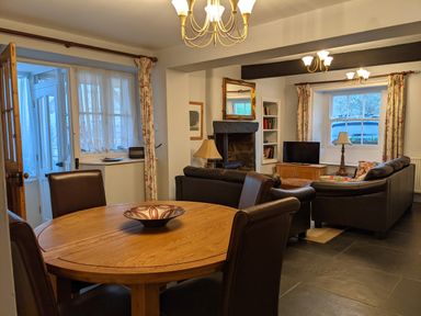 A dining area with a round wooden table and chairs next to a living room with sofas a fireplace and a television at Rose Cottage in Lowick Bridge