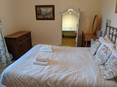 A bedroom with a bed arranged with white linens and folded towels a wooden chest of drawers a dressing table with a round mirror and a doorway leading to a bathroom at Rose Cottage in Lowick Bridge
