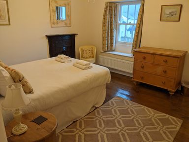 A bedroom with a bed, a wooden dresser, a chair, a side table with a lamp, and a window with curtains at Rose Cottage in Lowick Bridge