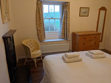 A bedroom with a bed and towels on it a wooden dresser a chair near a window with curtains and a framed picture on the wall at Rose Cottage in Lowick Bridge