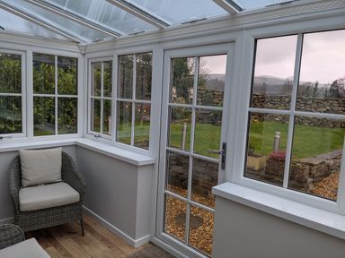 A conservatory with a wicker chair and glass doors overlooking a garden with stone walls at Rose Cottage in Lowick Bridge