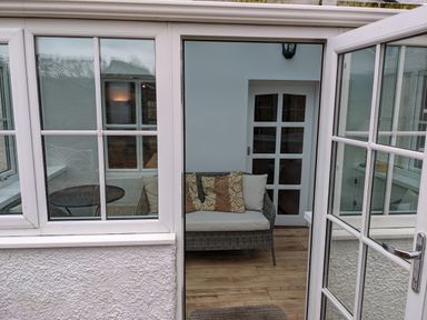 A small indoor seating area with a wicker loveseat and cushions next to a white door with glass panels at Rose Cottage in Lowick Bridge
