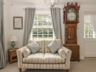 A sitting room with a striped sofa cushions a grandfather clock lamp on a wooden side table and curtains at Humble Bee Cottage in Wold Newton near Hunmanby