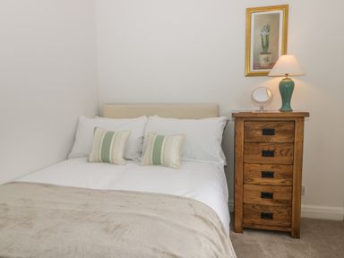 A bedroom with a bed with pillows and a blanket next to a wooden chest of drawers with a lamp and framed picture on the wall at Humble Bee Cottage in Wold Newton near Hunmanby