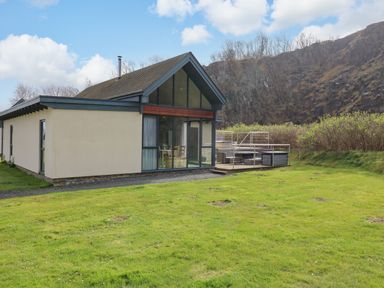 A house with large windows and a wooden deck with outdoor furniture on a grassy lawn near rocky hills at Restharrow Lodge in Bamburgh