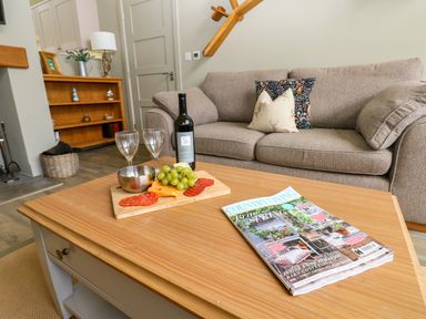 A living room with a beige sofa two decorative cushions a wooden coffee table with a magazine a bottle of wine two wine glasses grapes and cheese at Restharrow Lodge Bamburgh