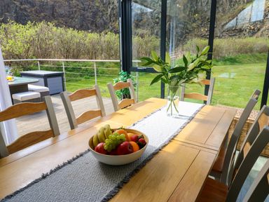 A dining table with a fruit bowl and flowers in a glass-walled room overlooking a garden at Restharrow Lodge Bamburgh