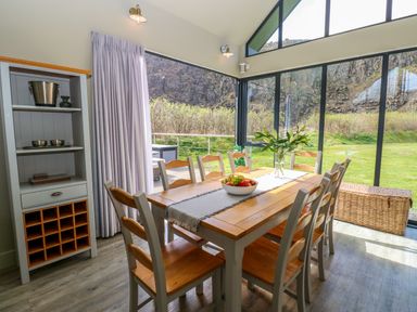 A dining area with a wooden table and chairs next to large glass windows showing an outdoor view at Restharrow Lodge in Bamburgh