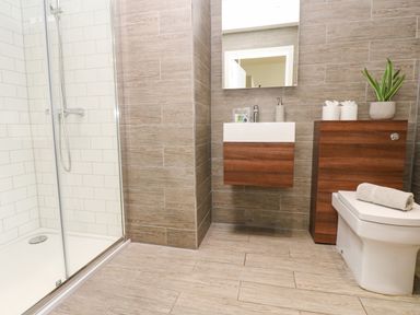 A bathroom with a shower, a toilet, a sink with a mirror, and wooden cabinets at Restharrow Lodge in Bamburgh
