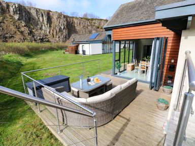 A patio with outdoor seating and a glass table next to a house with open folding doors and a grassy area with rocky cliffs in the background at Restharrow Lodge in Bamburgh