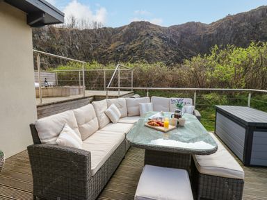 An outdoor deck area with a cushioned wicker sofa set and a glass table with breakfast items at Restharrow Lodge in Bamburgh