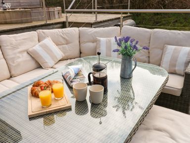 An outdoor seating area with a glass table two cups a French press a vase with purple flowers two glasses of orange juice and croissants at Restharrow Lodge Bamburgh
