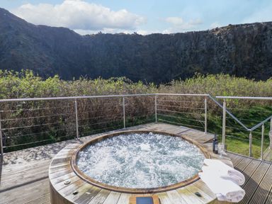 An outdoor wooden hot tub with bubbling water on a deck surrounded by metal railings with a mountain and bushes in the background at Restharrow Lodge in Bamburgh