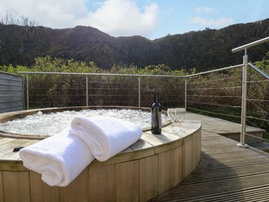 An outdoor wooden hot tub with white towels a bottle and two glasses on the edge with hills and bushes in the background at Restharrow Lodge in Bamburgh