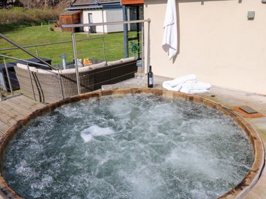 A bubbling outdoor hot tub on a wooden deck with towels bottle and glasses near a wicker sofa at Restharrow Lodge in Bamburgh