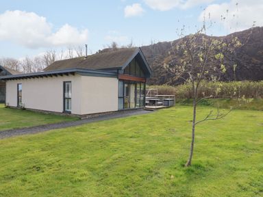A modern single story house with large windows on a grassy lawn with a small tree and hills in the background at Restharrow Lodge in Bamburgh