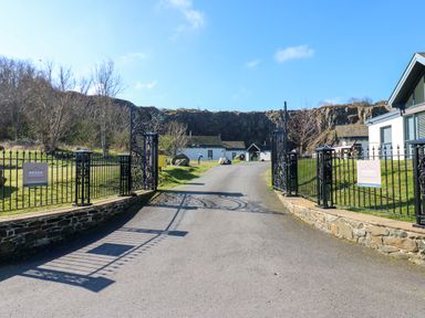The entrance gate to a property with a paved driveway leading to white buildings with a rocky hill in the background at Restharrow Lodge in Bamburgh