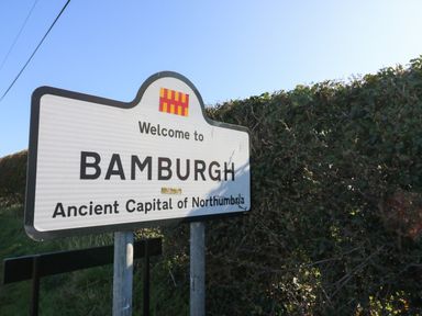 A welcome sign for Bamburgh beside a hedge under a clear sky