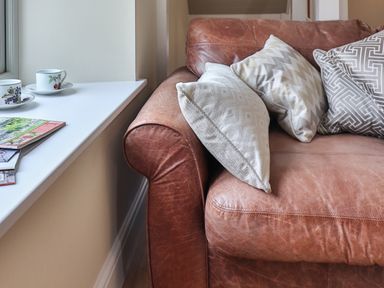 A brown leather sofa with patterned cushions next to a white windowsill with teacups and magazines at The Retreat in Lympsham