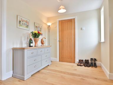 A hallway with a dresser holding a vase of flowers and a bottle of wine next to a wooden door with boots on the floor at The Retreat in Lympsham