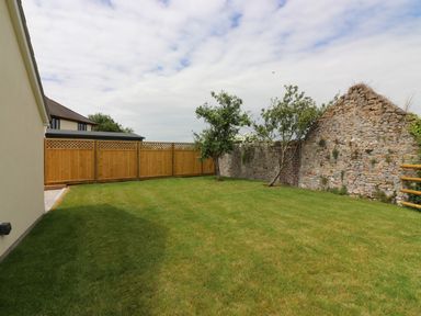 A fenced backyard with grass and two small trees next to a stone wall at The Retreat in Lympsham