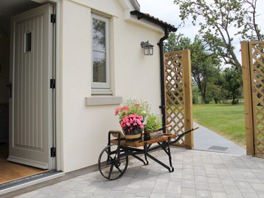 A wooden wheelbarrow with potted flowers near an open door and garden gate at The Retreat in Lympsham