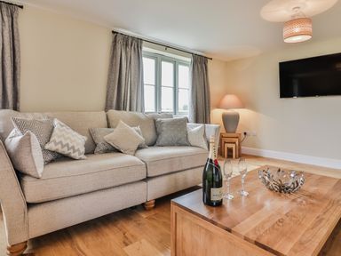 A living room with a beige sofa with patterned cushions a wooden coffee table with a bottle of champagne and glasses a table lamp and a wall-mounted television at The Retreat in Lympsham