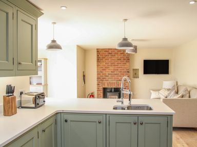 A kitchen with green cabinets and a countertop with a sink toaster and knife block near a living room with a brick fireplace and a mounted TV at The Retreat in Lympsham