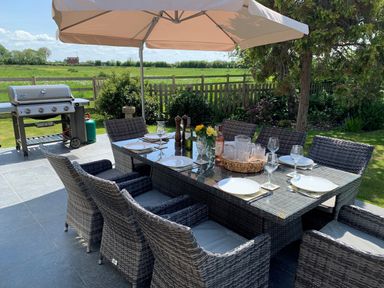 An outdoor patio dining set with a glass table and wicker chairs under a large umbrella next to a barbecue grill in a garden at The Retreat in Lympsham