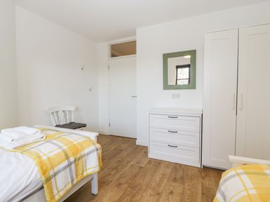 A bedroom with two single beds with yellow plaid blankets a white chair a white chest of drawers with a green framed mirror and a white wardrobe at Stable Cottage in Shipham
