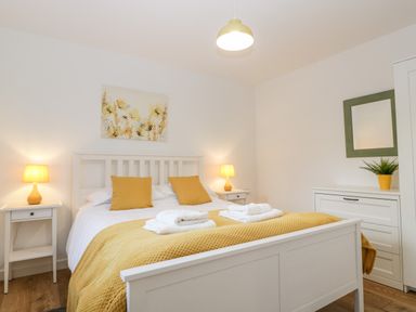 A bedroom with a white bed frame yellow pillows and a yellow bedspread bedside tables with lamps a dresser with a plant and mirror at Stable Cottage in Shipham