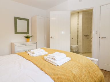 A bedroom with a yellow blanket on the bed white towels on the bed a white dresser with a yellow potted plant a green framed mirror a white wardrobe and a bathroom with tiled walls at Stable Cottage in Shipham