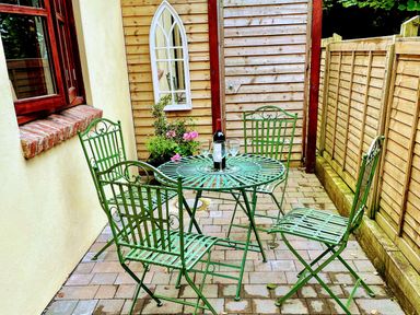 A patio area with a green metal table and four chairs with a bottle and glasses on the table and flowers near a wooden shed at Stable Cottage in Shipham