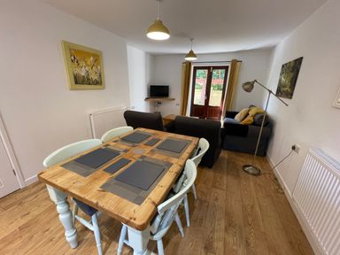 A dining table with four chairs and placemats in a room with sofas a floor lamp and a television at Stable Cottage in Shipham