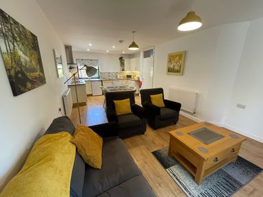 A living room with a sofa and armchairs around a coffee table next to an open kitchen with dining table at Stable Cottage in Shipham