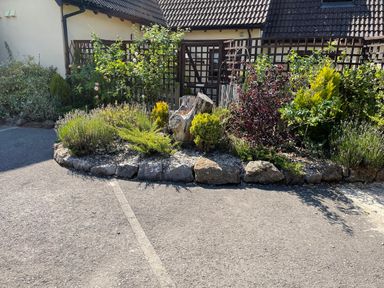 A garden bed with plants and a tree stump bordered by rocks in front of a house with a tiled roof at Stable Cottage in Shipham