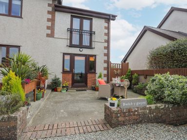 A patio area with chairs and a table outside a two-story house with plants and a sign reading Bwlch y gwynt at Bwlch Y Gwynt Bach in Llangefni