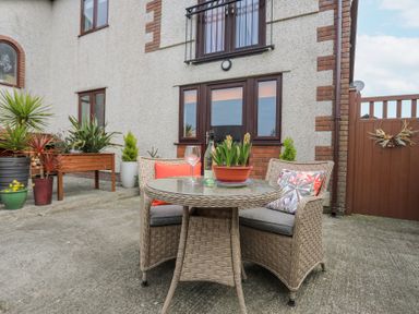 An outdoor patio area with a round wicker table and two wicker chairs with cushions and pillows in front of a house with potted plants and a wooden gate at Bwlch Y Gwynt Bach in Llangefni