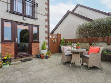 A patio with a round table and two chairs with cushions and plants in pots near a house at Bwlch Y Gwynt Bach in Llangefni
